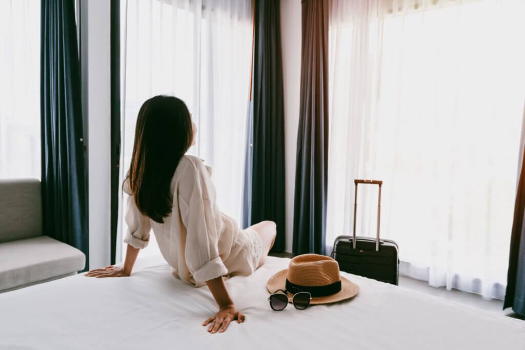 Young asian woman traveler with luggage sitting and relaxing on the bed in hotel room - Midas Travel Management Support your corporate travellers with their mental health